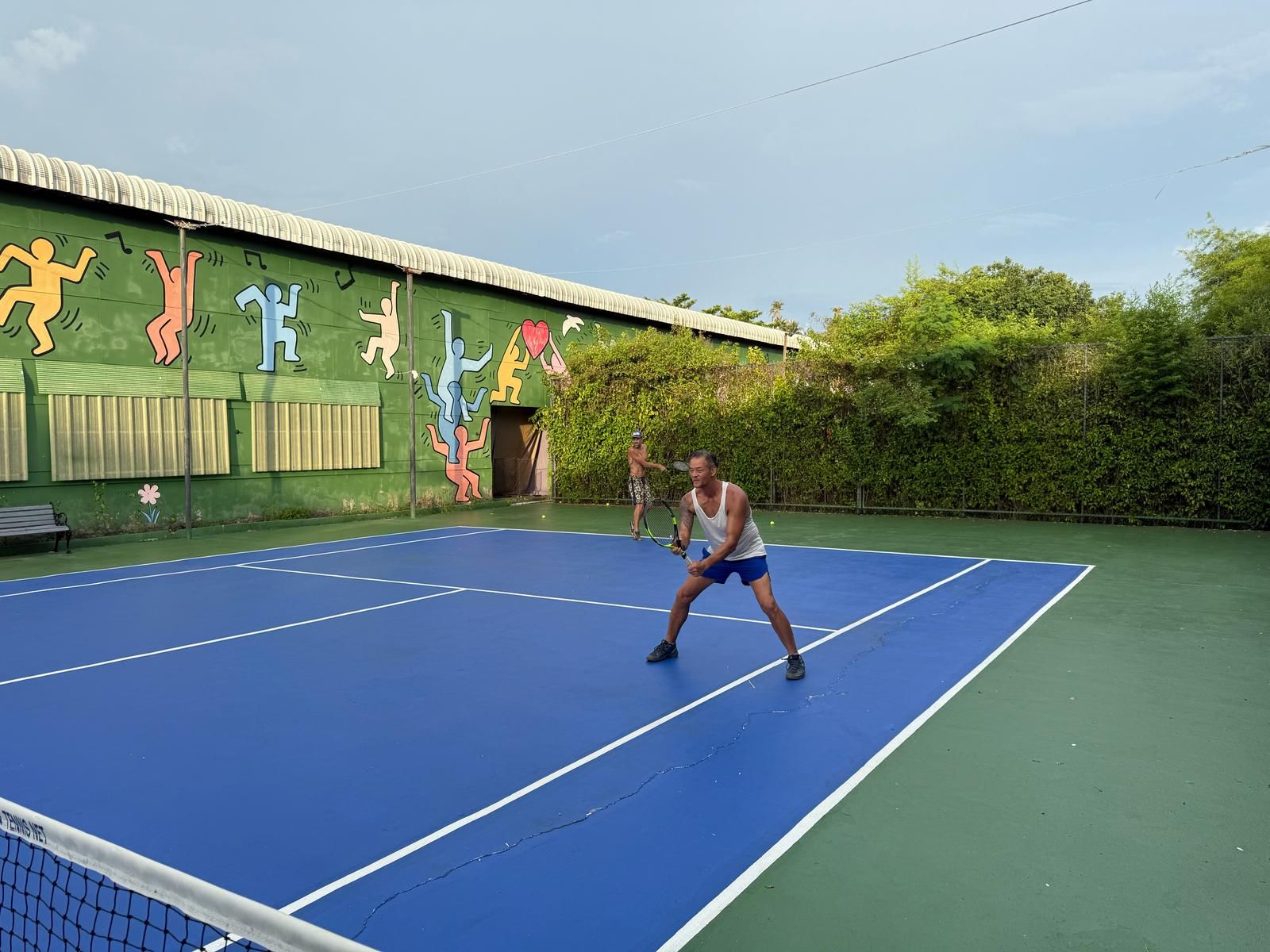 Tennis players rallying on an open-air blue hard court with a colorful mural backdrop on Koh Mak