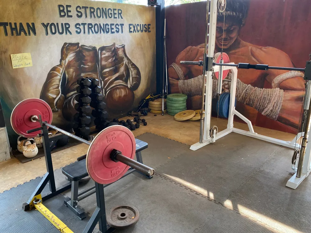 The strength corner of a Muay Thai gym on Koh Mak, with a loaded barbell on a bench, dumbbells on an A-frame rack and kettlebells beside painted boxing-glove murals