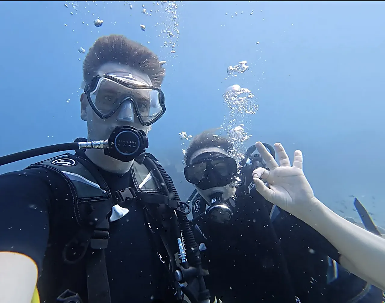 Two scuba divers below the surface near Koh Mak, one giving the OK hand signal as bubbles rise towards the sunlight