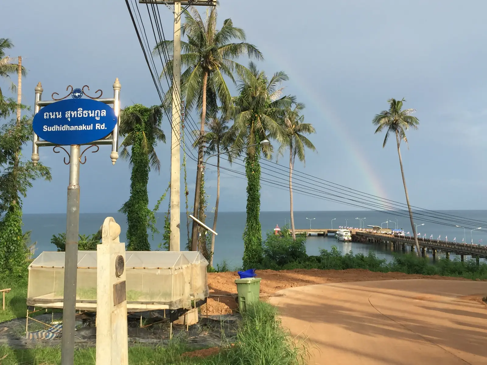 Koh Mak's main road with Sudhidhanakul Road sign leading toward Ao Nid Pier and a rainbow over the sea