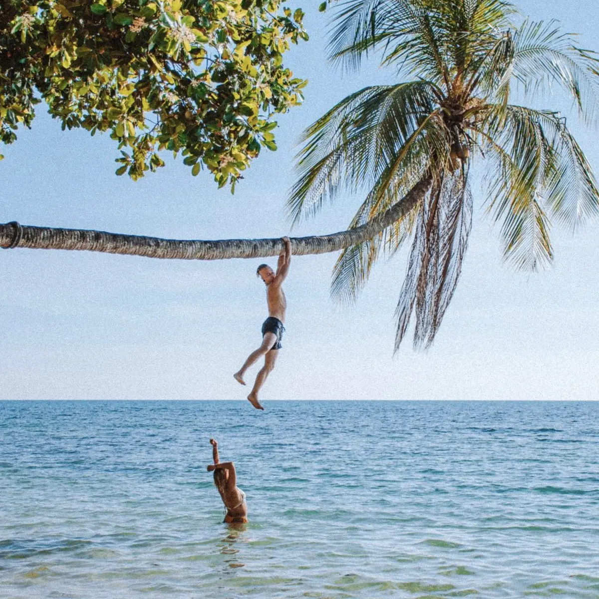 Two people playing on a leaning palm tree over calm turquoise sea at Ao Kao Beach, Koh Mak