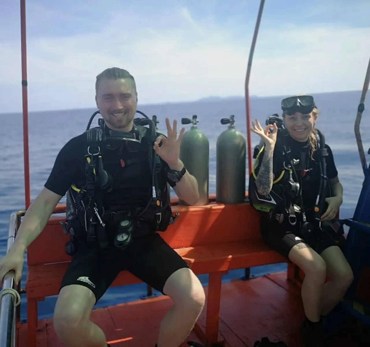 Two scuba divers in full gear sitting side by side on the deck of a dive boat, both giving the OK hand signal